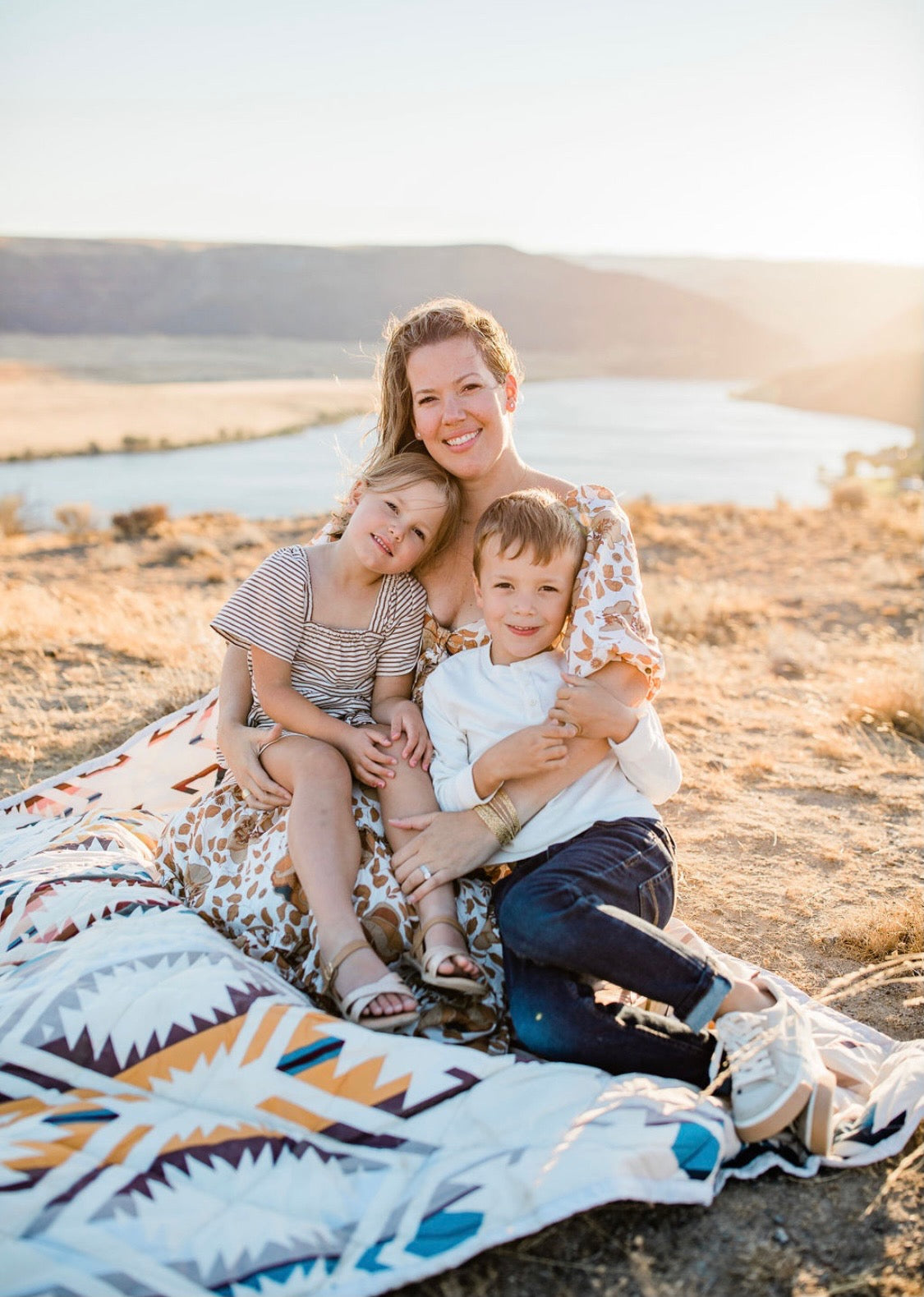 A mother and two children sitting together outdoors on a patterned blanket
